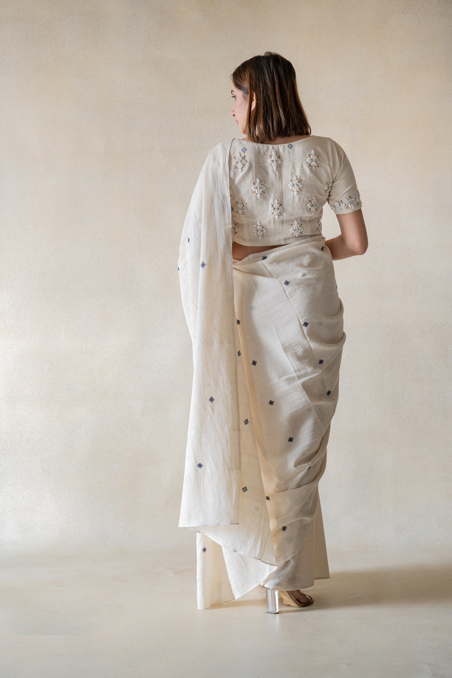 Experience the delicate grace of our off-white, handspun organic cotton saree and embroidered blouse ensemble. Slowly crafted with artful detail, this set features a beautiful booti design and mesmerizing hand embroidery on the sleeves and back. Its closed round neck with hook and eye closure exudes a delightful and calm aura.
