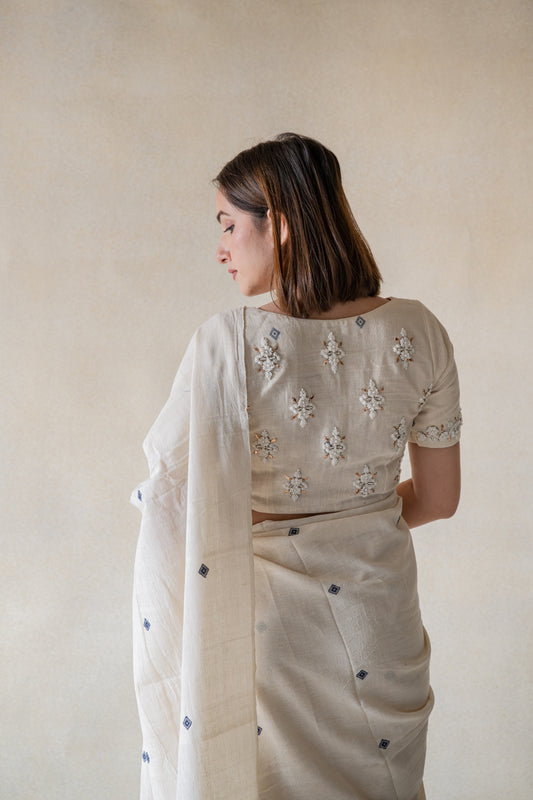 Experience the delicate grace of our off-white, handspun organic cotton saree and embroidered blouse ensemble. Slowly crafted with artful detail, this set features a beautiful booti design and mesmerizing hand embroidery on the sleeves and back. Its closed round neck with hook and eye closure exudes a delightful and calm aura.